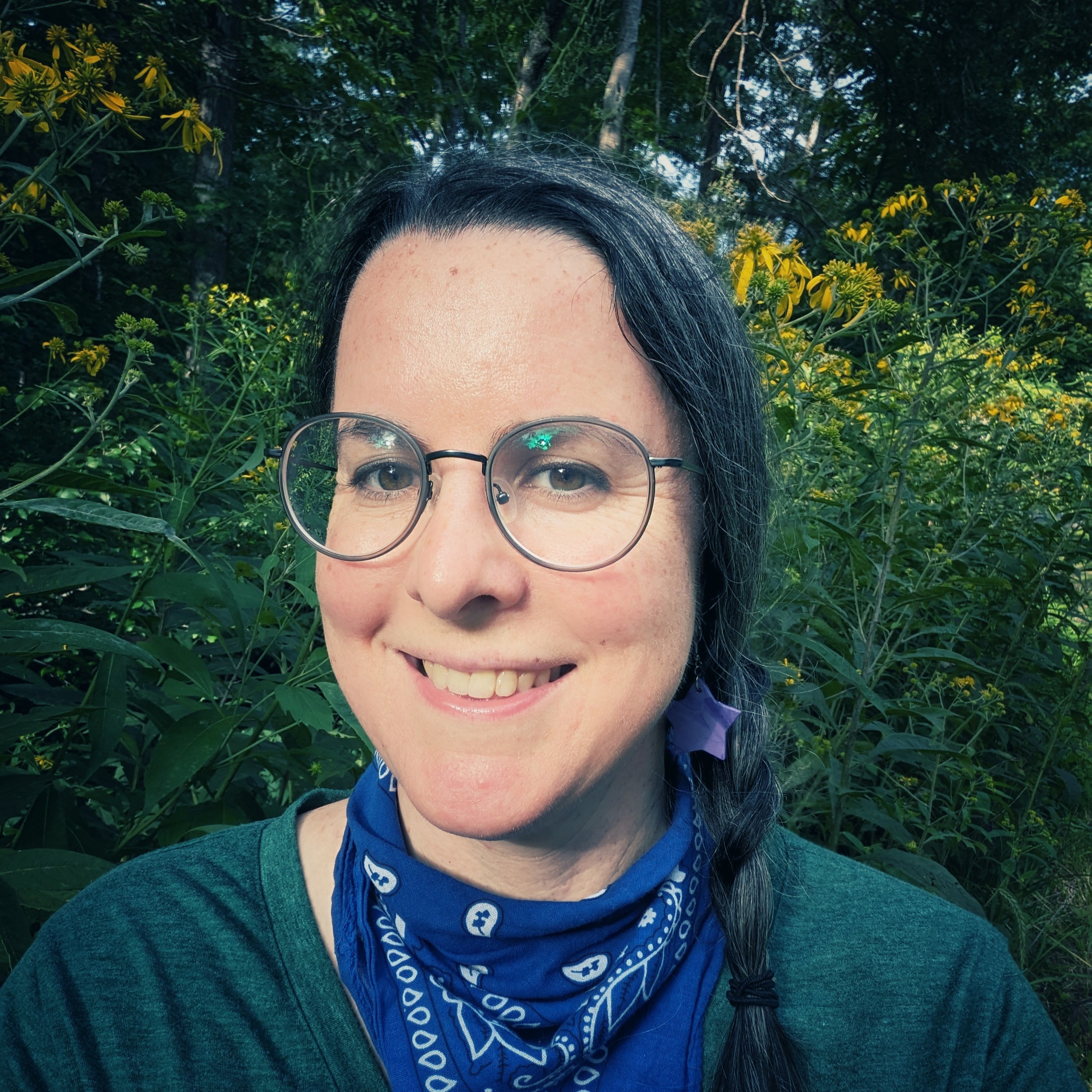 Cassie wearing green, blue, and glasses with a background of forest and yellow flowers